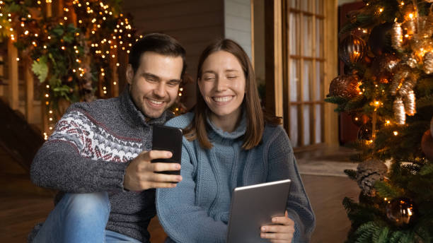 istock Happy bonding young couple using gadgets, sitting near Christmas tree. 1695600813