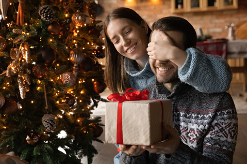 Happy beautiful millennial woman covering eyes of curious young husband, presenting Christmas gift, making surprise at New Year night. Joyful sincere couple celebrating winter holidays together.
