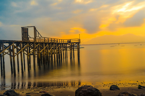 Wooden pier juts out into calm body of water, bathed in golden light of sunrise in Banyuwangi. Peaceful scene invites reflection and relaxation