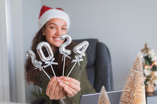 Cheerful young business woman celebrating new year at work, holding balloons shaped as 2024 numbers and wearing a Santa Claus hat