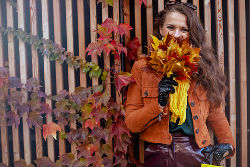 Hello october. smiling elegant middle aged woman in brown trench coat with autumn yellow leaves in the city.