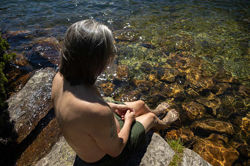 An adult man sits on the shore of a mountain lake and cools his feet in the water
