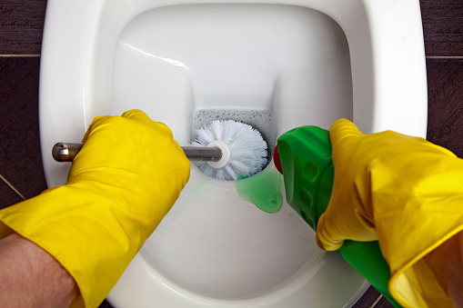 The hands of a person in yellow rubber gloves using a disinfectant gel and a toilet brush wash the toilet.