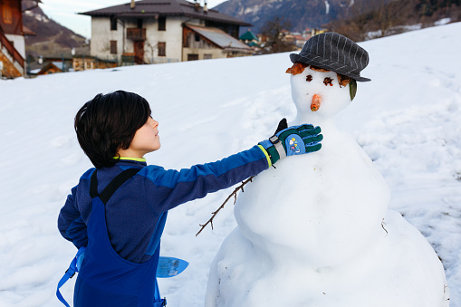child with short dark hair in blue winter clothes making snow man on snow slope