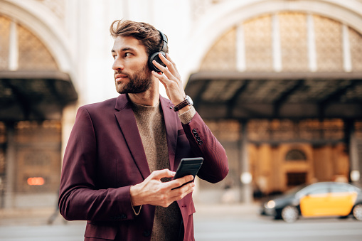 Handsome businessman walks down the street and listens to music with headphones. He is happy and smiled. Positive city life concept.
