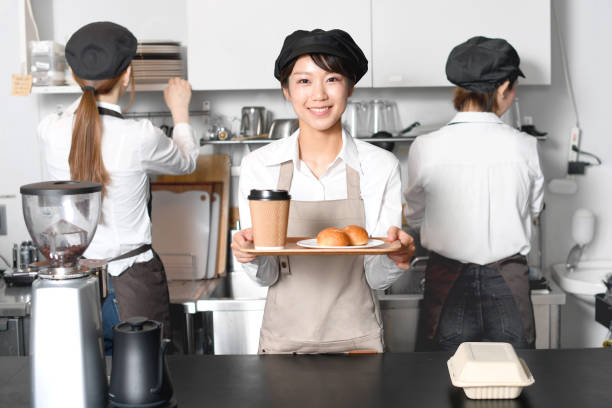 식당의 주방 카운터에서 미소를 지으며 손님을 접대하는 젊은 아시아 여성과 주방에서 일하는 젊은 여성 직원. - soda jerk 뉴스 사진 이미지