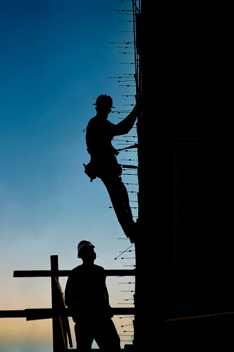 Vertical silhouette of a construction worker in harness on the side of a building under construction with sunset background