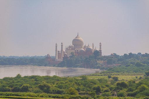 beautiful white marble Taj Mahal is seen from the Red Fort . In the foreground are green fields, a river,  blue sky. India, Agra.
