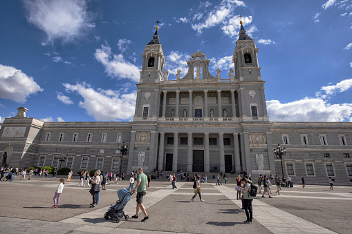 Exterior view of Almudena Cathedral on a sunny afternoon, Madrid, Spain. Many tourists at the Plaza de Armeria can be seen.