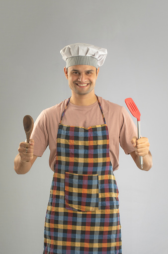 Portrait of smiling male chef dressed in hat and apron holding wooden spoon and spatula while standing on white background