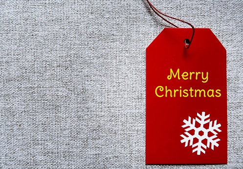 Red MERRY CHRISTMAS gift tag on gray fabric background with copy space - concept of Christmas holiday season, festive joyful celebration , present giving and warm wishes to love ones