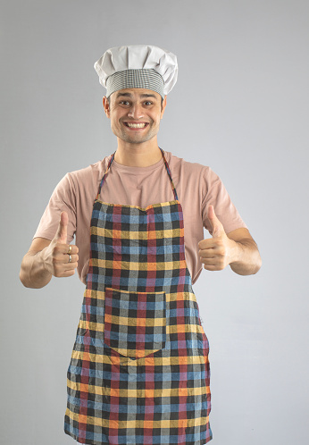 Portrait of confident smiling male chef wearing hat and apron showing thumbs up gesture against white background