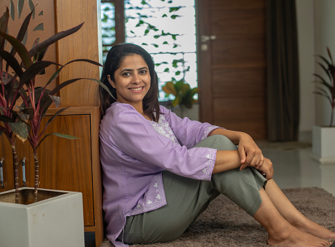 Portrait of smiling beautiful woman hugging her knees and sitting on carpet by wooden closet and potted plant at home