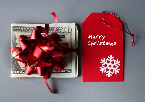 Cash dollars money in a red ribbon and gift bow and red Christmas gift tag with written text MERRY CHRISTMAS, concept of money bonus reward giving to employee or family love ones