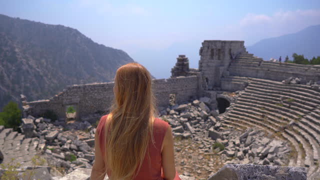 A young woman embarks on a cultural journey as she explores the breathtaking ruins of Termessos, nestled within the mountains near Antalya, Turkey. She sits in on the steps of an ancient amphitheater embracing the spirit of this site. This footage invite
