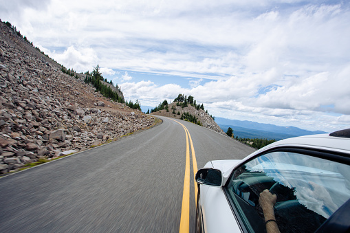 Road trip image of a white car traveling along a mountain pass road in the back country of Oregon's Crater Lake National park. This image shows the driver's hands on the steering wheel, and has intentional motion blur from the movement and speed of the car. Road is a single lane, two way paved road with a turn ahead.