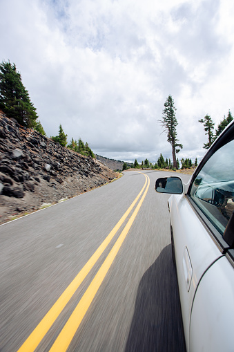 Road trip image of a white car traveling along a mountain pass road in the back country of Oregon's Crater Lake National park. This image shows the driver's hands on the steering wheel, and has intentional motion blur from the movement and speed of the car. Road is a single lane, two way paved road with a turn ahead.