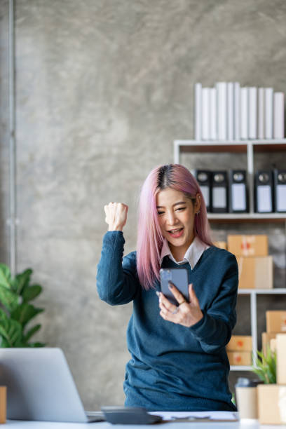 Portrait of an Asian businesswoman, a small business owner who successfully sells online and happily holds a smartphone with a laptop computer. Portrait of an Asian businesswoman, a small business owner who successfully sells online and happily holds a smartphone with a laptop computer. newspaper seller stock pictures, royalty-free photos & images