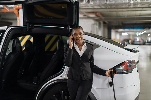 African female listening to phone caller whilt filling up electric motorcar from EV charger in basement garage. Efficient worker in formal wear reaping benefits of parking time in office building.