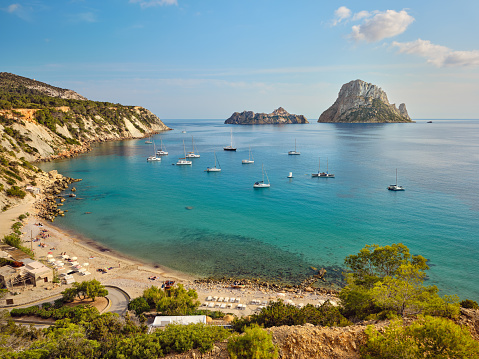 Wide-angle view of the bay of Cala d'Hort, on the southern coast of Ibiza, renowned for its crystal-clear waters and its stunning view on the iconic rocky isles of Es Vedrà and Es Vedranell. The warm light of a Mediterranean summer afternoon, picturesque clouds, sailing boats rocking gently on transparent waters, colourful cliffs covered with lush pine trees and Mediterranean scrub, bathers engaged in different activities. High level of detail, natural rendition, realistic feel. Developed from RAW.