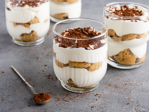 Traditional coffee, lady fingers biscuits, whipped cream and mascarpone trifles decorated with cocoa powder and served in drinking glasses with tea spoon.
