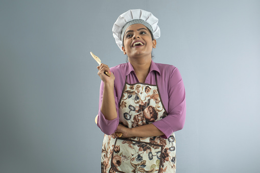 Portrait of smiling female chef dressed in hat and apron holding wooden spoon and spatula while standing on white background