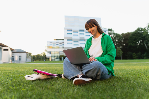 Young woman engrossed in her studies, seated on the grass with her laptop, set against the scenic backdrop of the campus, embodying a serene scholarly atmosphere
