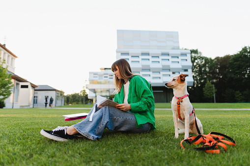 Studious female student, surrounded by the campus landscape, diligently takes notes while her loyal dog keeps her company, creating a charming scene of academic dedication and companionship