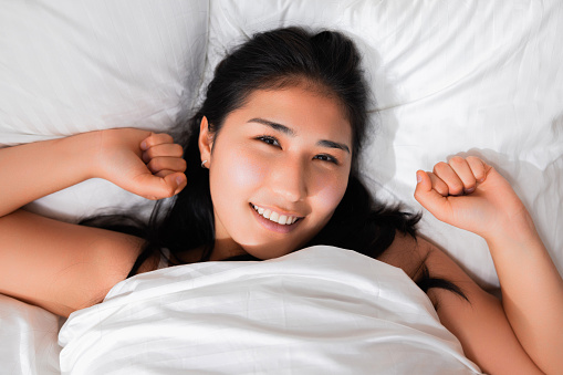 Cheerful adorable young japanese chinese kazachstan girl with long dark hair laying in bed top view side good morning concept. Delighted girl waking up at weekends smiling.