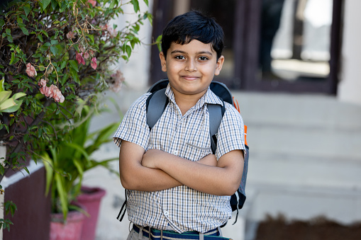 Portrait of happy indian boy student wearing school backpack standing cross arms outside the primary school looking at camera. Education concept.