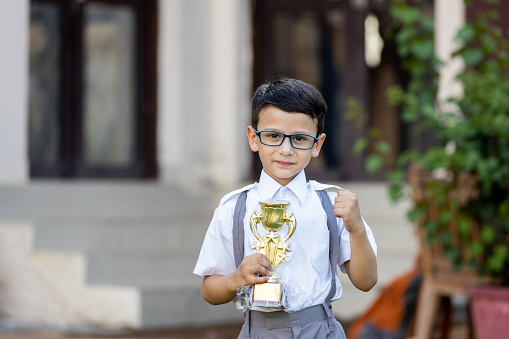 Happy Indian student schoolboy wearing school uniform holding victory trophy in hand outside school. Education concept.