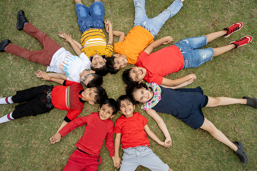Group of happy Indian kids wearing colorful cloths lying on grass in the summer park, Playful asian children playing together at outdoor garden. View from above. Background. Looking at camera.