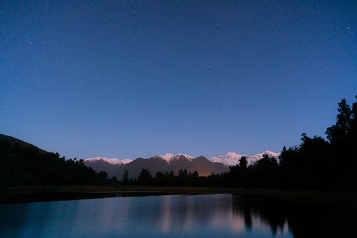 As evening sets in, stars appear overhead and are reflected in the mirror-like surface of spectacular Lake Matheson on New Zealand’s South Island. Beyond we see the snow capped peaks of Mt Tasman and Mt Cook and the Southern Alps.