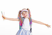 Asorable schoolgir with colorful braids and tennis cap stands on white background isolated holding paint brushes in hand wearing stylish clothes delightfully look at camera.Back to school concept