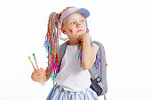 Adorable cute kid goes in frist form holds painting brushea in hands wears tennis cap with colorful braids standing on white background posing with beautiful small backpack