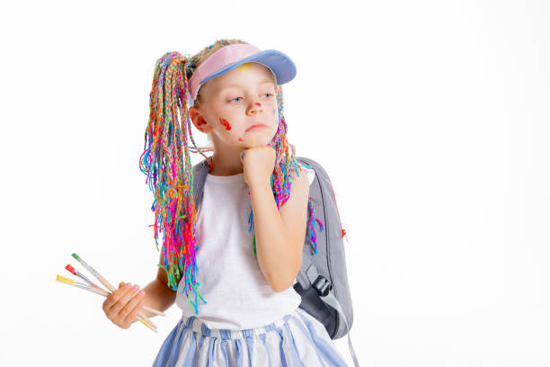 Beautiful small kid pupil shows sad emotions doesn't want to go to school first experience stands on white background with colorful braids wears rucksack backpack holds painting brushes.