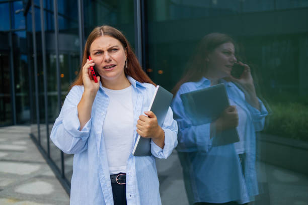 istock Serious Caucasian student freelancer blonde woman rest use laptop pc computer talk by mobile cell phone look, sitting outdoors against blurred city building. She is keyboarding on laptop. Freelance work concept. 1696722086