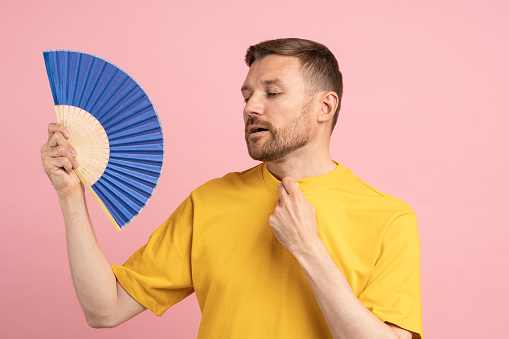 Sweaty exhausted middle aged man using paper fan suffer from heat feels sluggish. Displeased tired guy cooling in hot summer weather isolated on studio pink background. Overheating, stuffiness concept