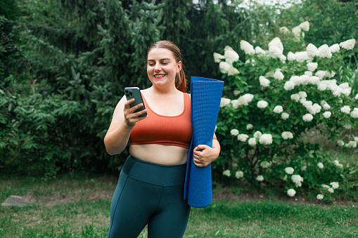 Young 20s fat woman hold yoga mat and using smartphone after workout on street. Portrait of smiling female in sportswear walk using smartphone. High quality photo