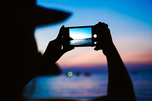 Woman taking photos of a sunset with a mobile phone. The concept of the photos on the phone. Close up the smartphone lies buried takes a photo of a bridge and sea in the background.