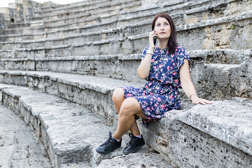 Mid adult woman is visiting the ruins of an ancient antique amphitheater. She is talking on her phone. The Ancient theatre of Ohrid of the Hellenistic period is located in Ohrid, North Macedonia.