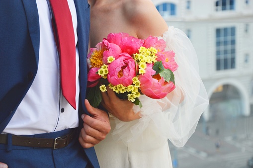 Wedding. The groom in a blue suit and the bride in a white dress stand side by side and hold a bouquet of bright pink flowers.
