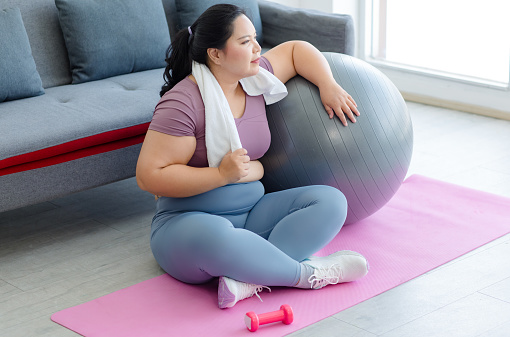 Asian young tired exhausted overweight oversized fat chubby plump female sportswoman in casual sportswear sitting on yoga mat wiping sweat with towel after working out with exercise ball and dumbbell