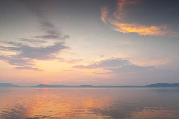Location: Lake Balaton, Hungary. As the sun gracefully descends on the horizon, its warm golden rays paint the sky in a mesmerizing palette of oranges, pinks, and purples. These hues gently reflect on the calm, glassy surface of the lake, creating a mirror-like effect that doubles the enchantment.