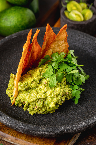 Avocado dip guacamole with crispy tortillas on black stone plate on wooden table