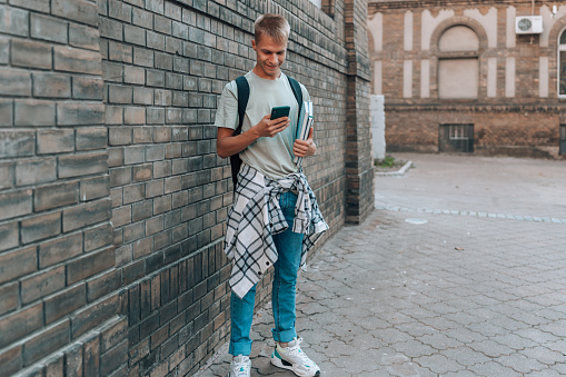 Standing before the impressive façade of the university, a young man is absorbed in the contents of his phone, disconnecting from his surroundings