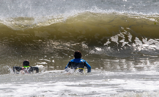 Gilgo Beach, New York, USA - 31 August 2023: Two men lying on their surfboards looking up at a very big wave.