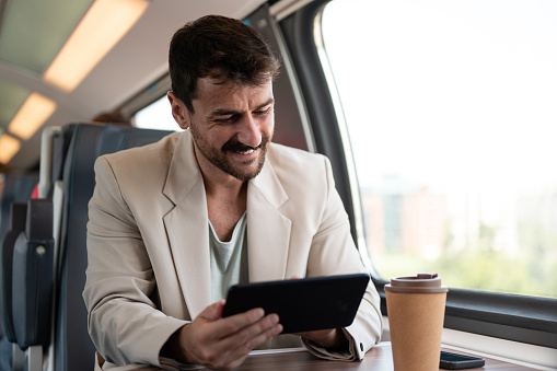 Young man enjoying his trip in train, using tablet and drinking coffee