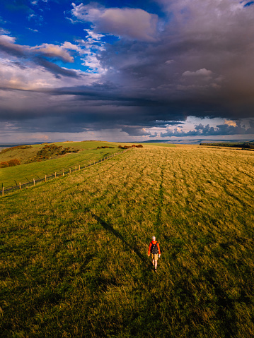 High angle image depicting one man hiking in the natural beauty of the South Downs in southeast England, UK. The man wears a red hoodie and black rucksack. It is golden hour and the land is bathed in soft warm light.