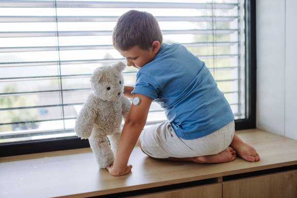 menino diabético com um monitor de glicose contínuo sentado à janela, mostrando seu sensor de ursinho de pelúcia no braço. - hypoglycemia diabetes child hyperglycemia - fotografias e filmes do acervo